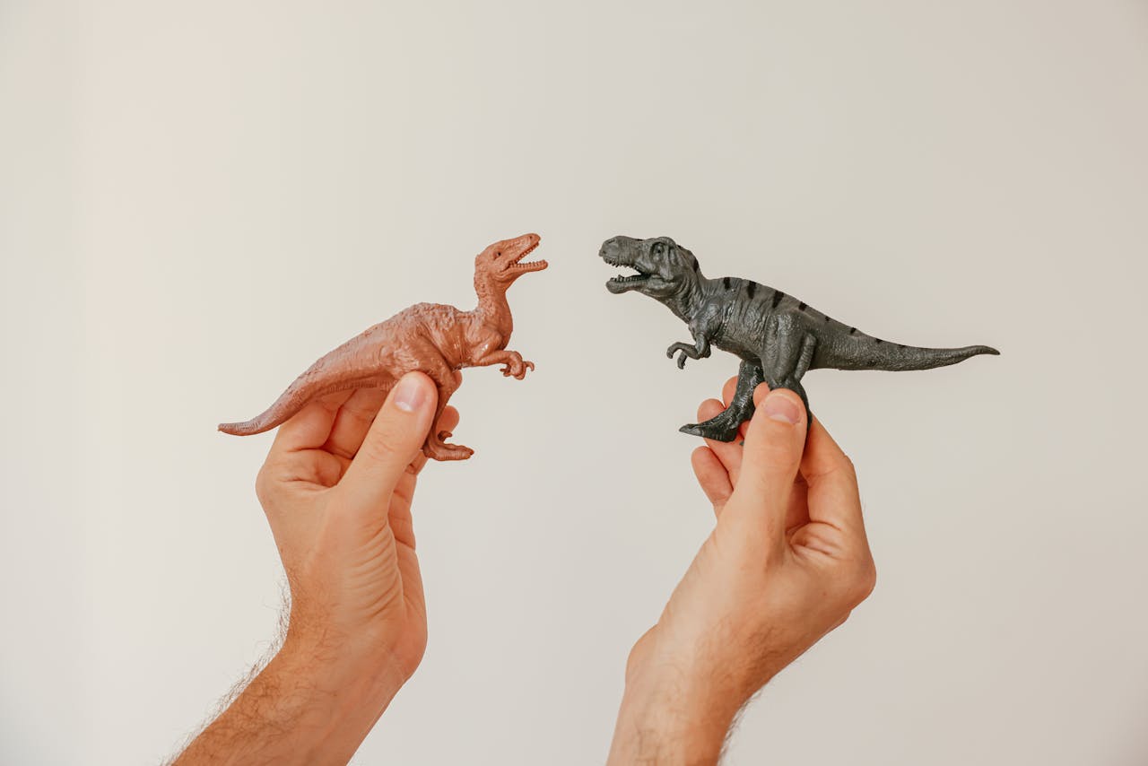 Hands hold toy dinosaurs playfully posed against a white background.
